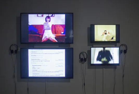 4 wall-mounted monitors, 1 show a young woman on a red sofa, 2 show young male figures and another displays text.
