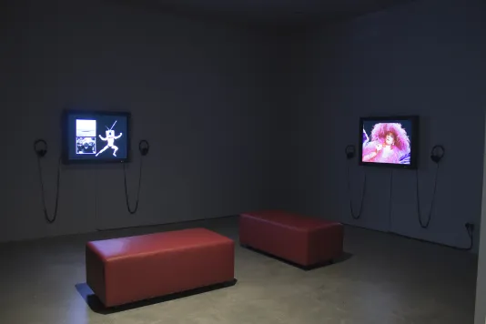 2 wall-mounted monitors with headphones show videos in a gallery with 2 red benches for visitors