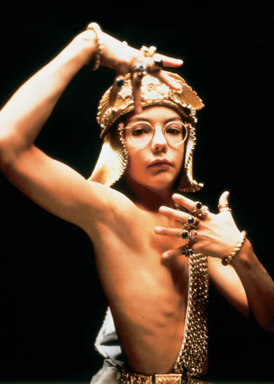 A shirtless boy costumed in gold head-dress, sash, rings, bracelets, and eyeglasses looks at the camera in a dance pose.