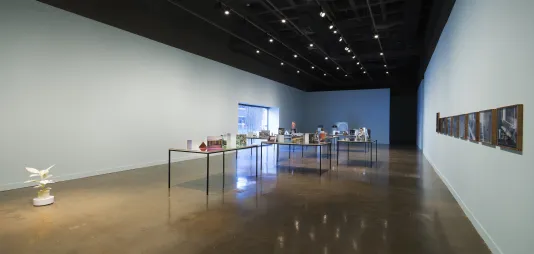 A gallery with several tables, a moving plant sculpture, pictures on the right wall, and a picture window to the outside