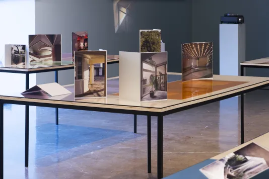 Installation view of tables with photographs arranged at varied angles, bathed in light from the picture window