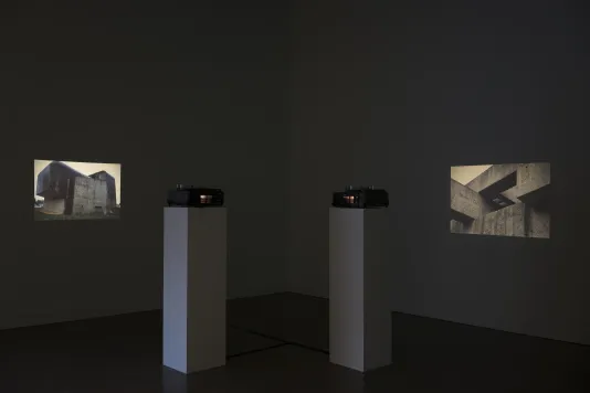 In a darkened space, two slide projections; on the left an image of a building and at right a cropped image of a structure