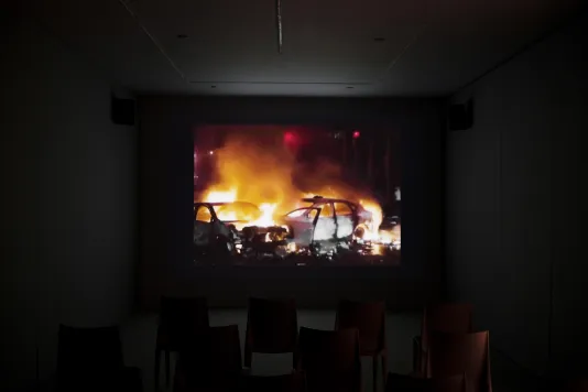 On a screen in a dark room, a video displays a night scene of two car skeletons in flames.
