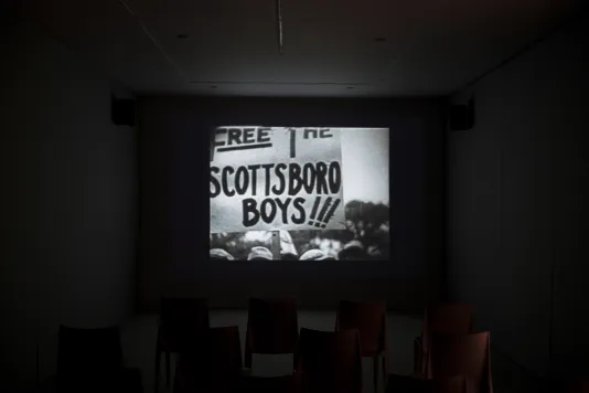 In the center of a dark room, a black and white video displays a protest sign that says “Free the Scottsboro Boys”.