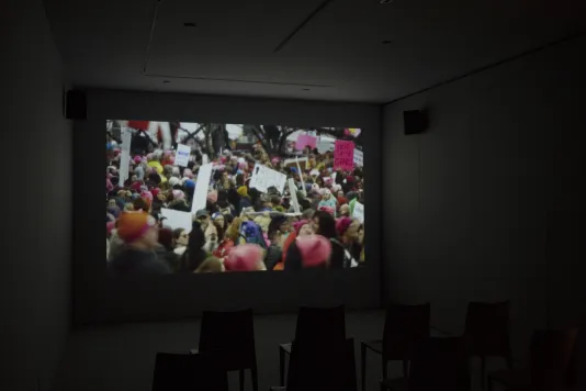 A colorful video scene of a large crowd of protesters, many holding signs, plays on a screen in a dark room.