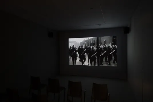 A black and white video of a group of police wearing helmets with face shields and clutching batons, displayed in a dark room.