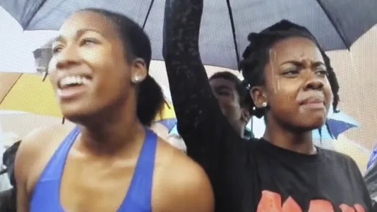 Closeup of two young women of color beneath an umbrella in a crowd, one is smiling broadly, the other has her arm raised.