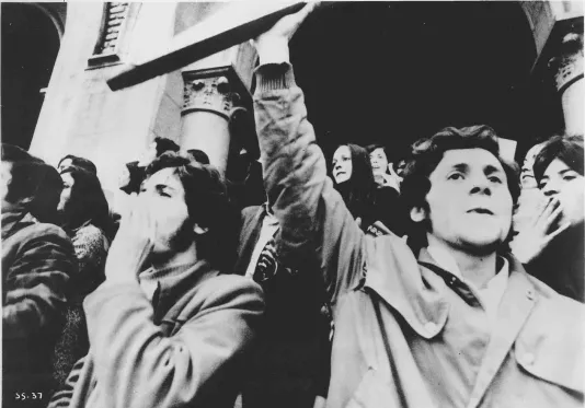 Black and white closeup still of college students protesting. A young man in front holds up an object cut off by the frame.