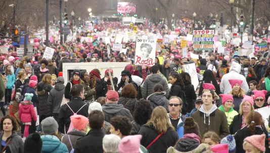 Still showing a massive crowd of protestors wearing pink pussy hats and holding signs, filling the screen.