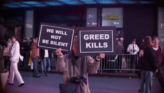A senior person holds 2 signs in front of their face, one reads We will not be silent, the other reads Greed Kills.