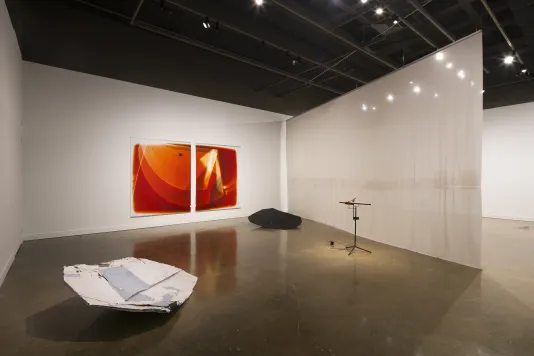 A large space bisected with hanging fabric, two large orange prints on a wall, a wooden sound piece, and two floor sculptures