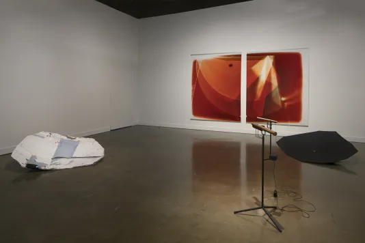 Two orange and white prints on a wall, a black and a white shield sculpture on the floor, a wood sound piece on a stand