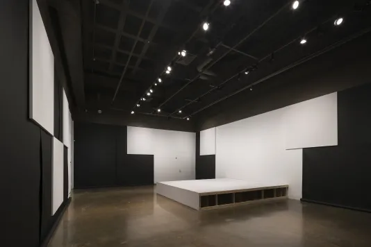 Three walls partially covered in black paper, white square panels overlapping the paper. A wooden stage abuts a wall at right.
