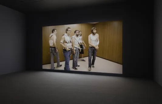 In a wood paneled space, five men looking toward the right, all wearing pants, tin foil vests, and belts with bells dangling