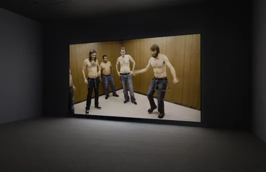 Four shirtless men, one mid-motion with arms outstretched, the other three watching, all wearing belts with bells dangling