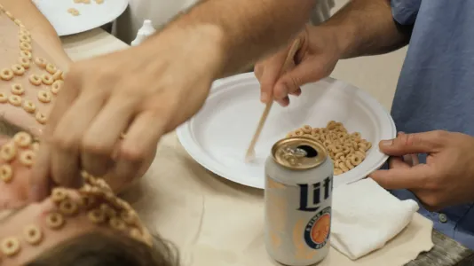 A can of Lite beer, a hand glueing Cheerios on another’s forehead, two hands holding a plate of Cheerios, and a glue stick