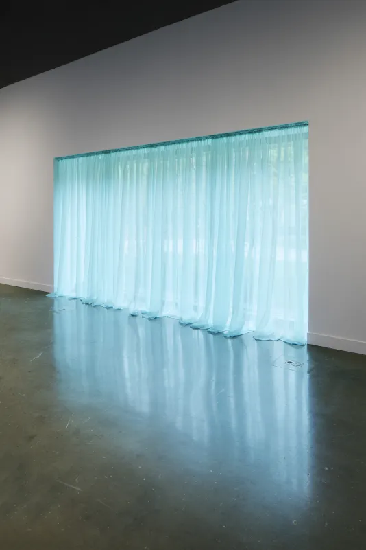 A light blue sheer curtain, the color of the sky, on a long, tall picture window along a white wall, shot on a diagonal