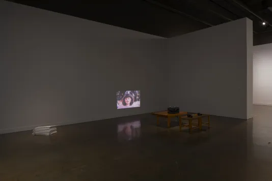 A dark space with a projection of a woman low on a wall, a projector on a small low table, a stack of linens on floor nearby 