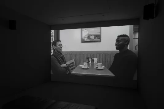 Black and white video projection of a woman and a man in conversation, sitting opposite at a cafe table set with coffee.