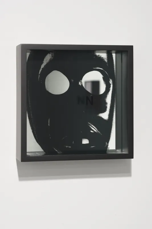 Framed close up image of a black mask, an eyehole reflects artwork from the opposite wall, the letter N sits on the glass.