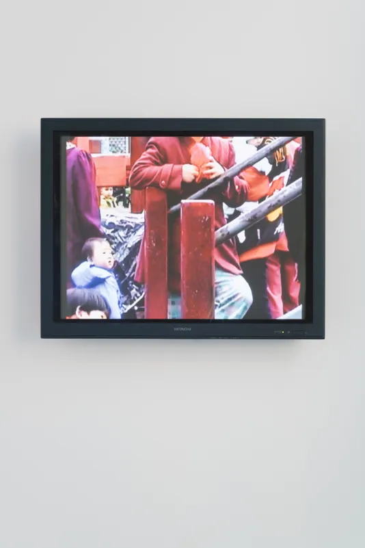 Video monitor shows a colorful scene of a torso in a red coat behind a railing, a small child, glimpses of others around them.