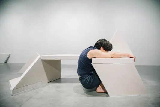 A person squats low with legs tucked under a bench sculpture, arms stretched over the top, and grasps the far edge of the seat.