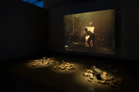 Three piles of gold-painted rocks and mallets are grouped in front of a screen on which an image of a man working in a forge.