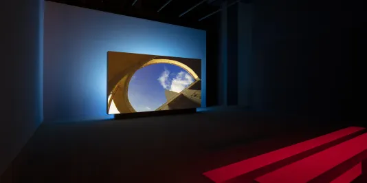 In a darkened gallery with red benches, a screen shows a projected image of blue sky seen through a roof opening.