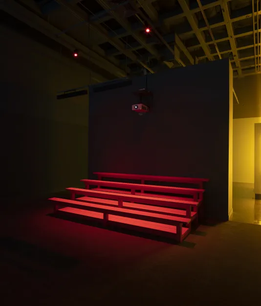 Red wooden bleachers are illuminated in an otherwise darkened Gallery. A yellow light is visible in a rear gallery.