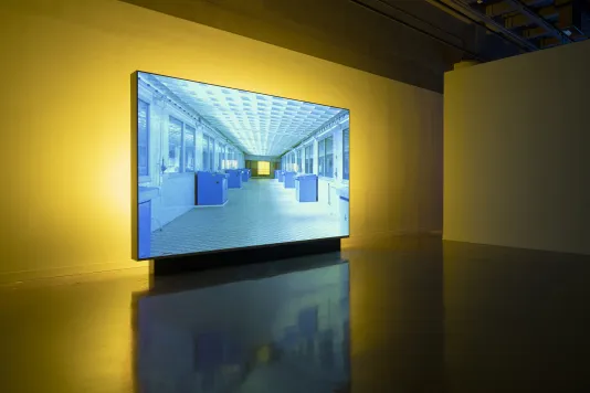 An image of a corridor with blue boxes and a patterned ceiling is projected on a standing screen backlit by yellow light.