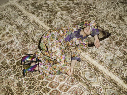 Woman in long floral-patterned dress reclines on and blends into the rich tan and red patterns of the carpet.
