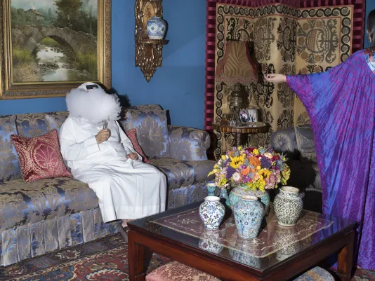 In a colorful ornately-patterned room, a puff of white smoke conceals the face of a man in white robe holding a silver pipe.