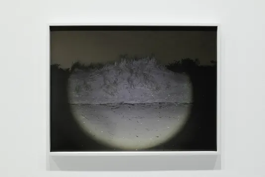 A semi-circle of light illuminates a sand dune against a sepia-colored sky in a framed photograph on a white gallery wall.