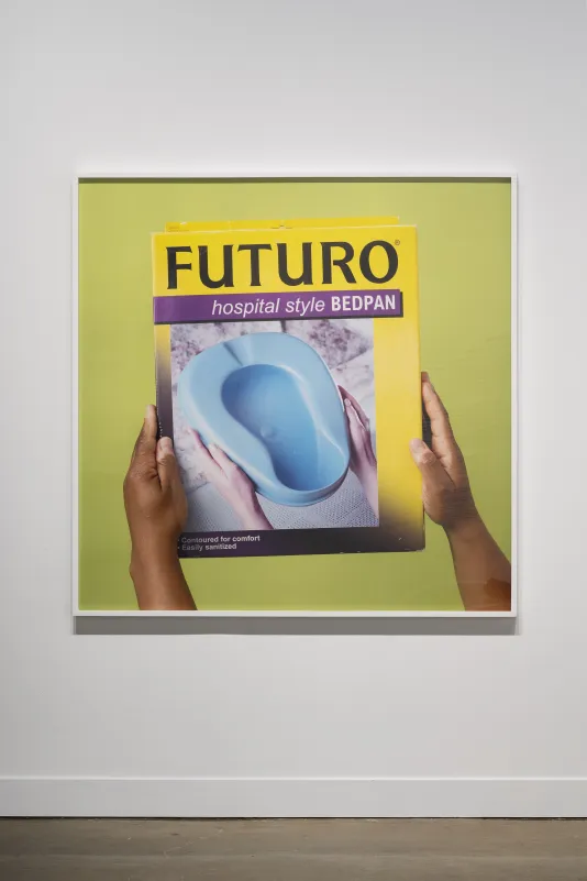Hands with dark skin tone hold a brightly colored box that reads: "Futuro Hospital Style Bedpan" and has an image of a light blue bedpan. 