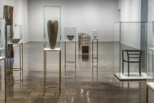 Vases and chairs seemingly made of concrete and metal sit atop bronze pedestals inside a glass case. Int he background are other works from Kwade's exhibition. 