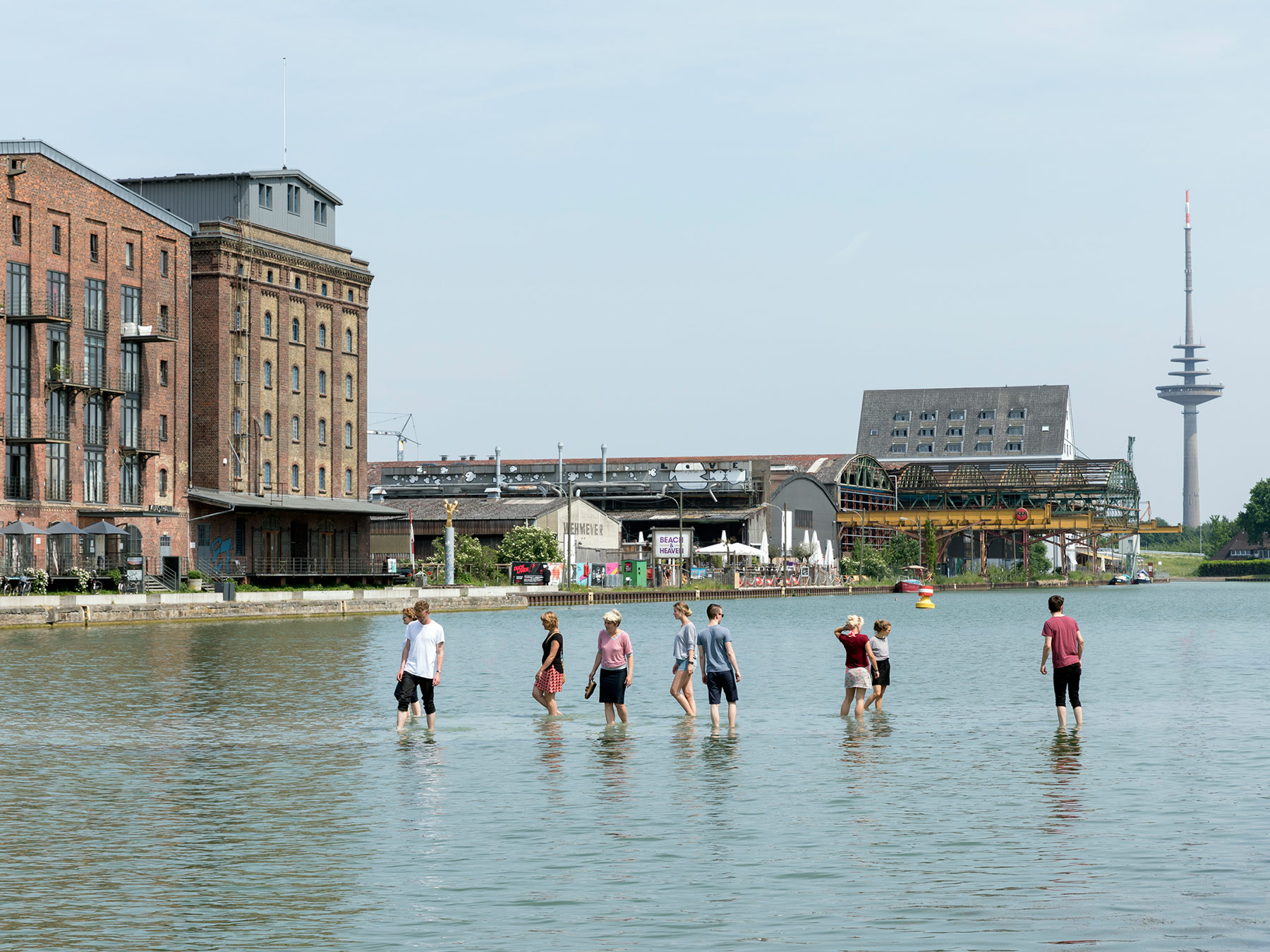 9 people walking in water up to their knees on a shore near buildings.