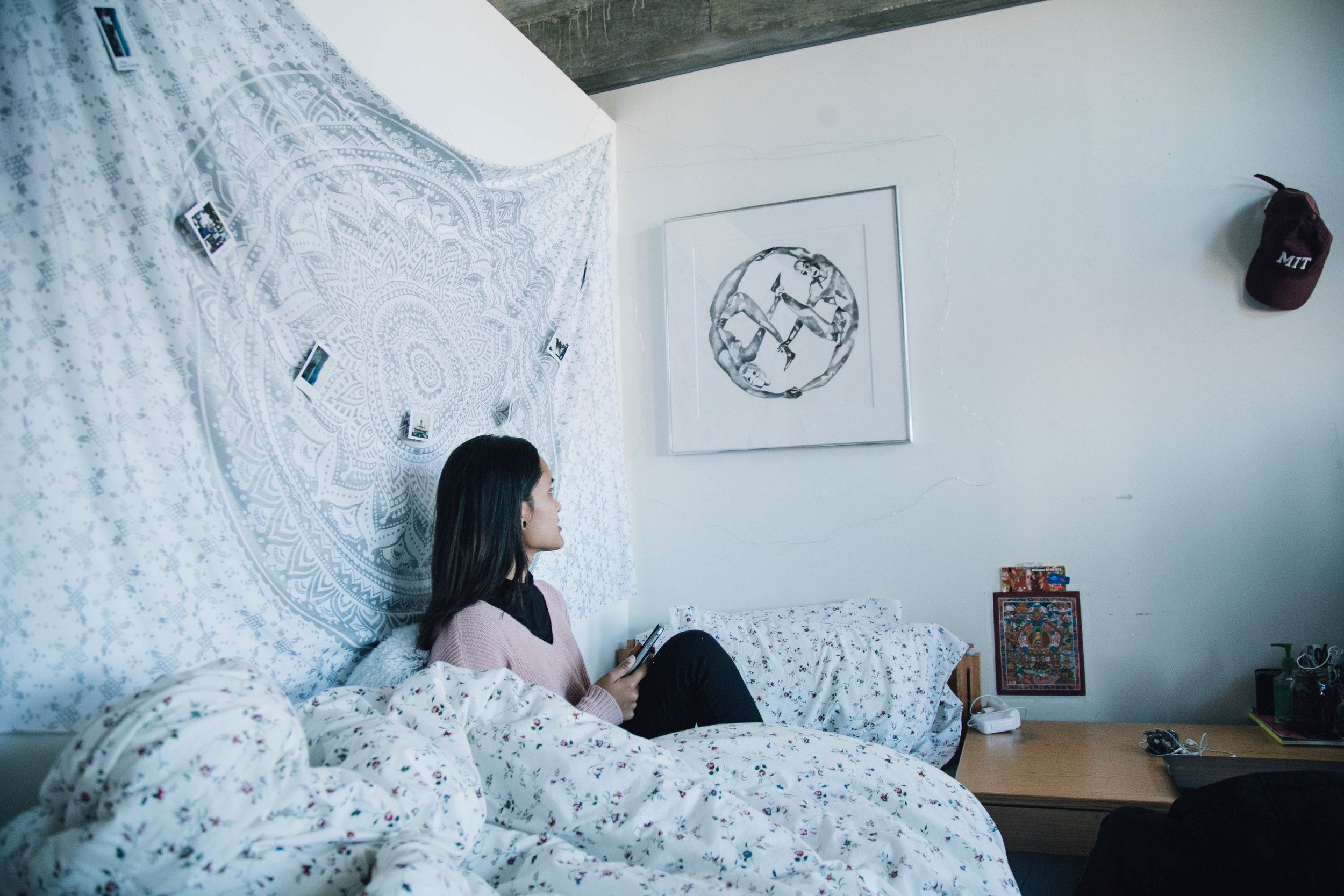 A woman sits on her dorm bed with a tapestry behind her gazing at a print on the wall to her left.