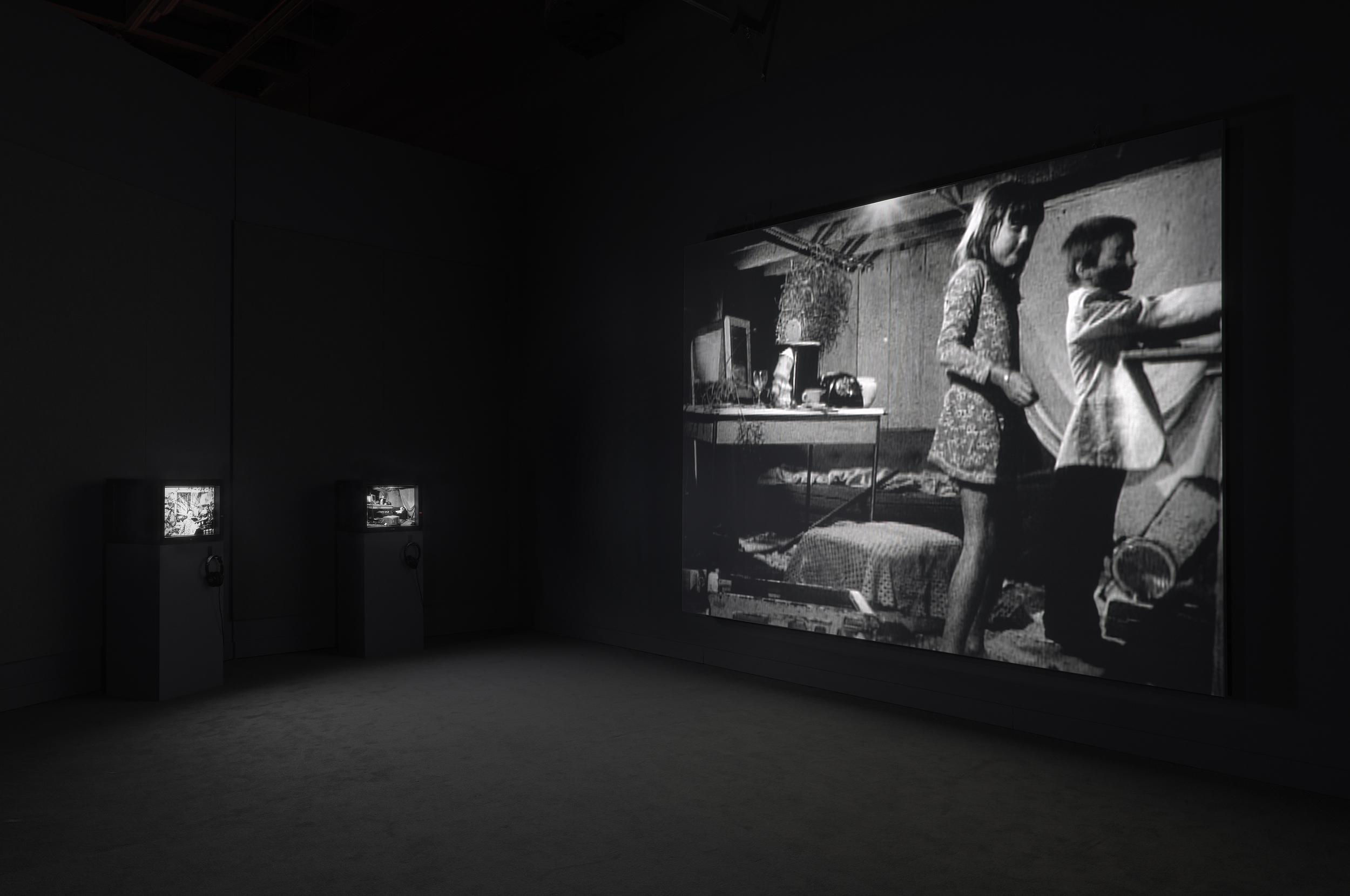 Black and white photo of video projection and three chairs. Projection features two children in a cluttered room with a desk and computer.