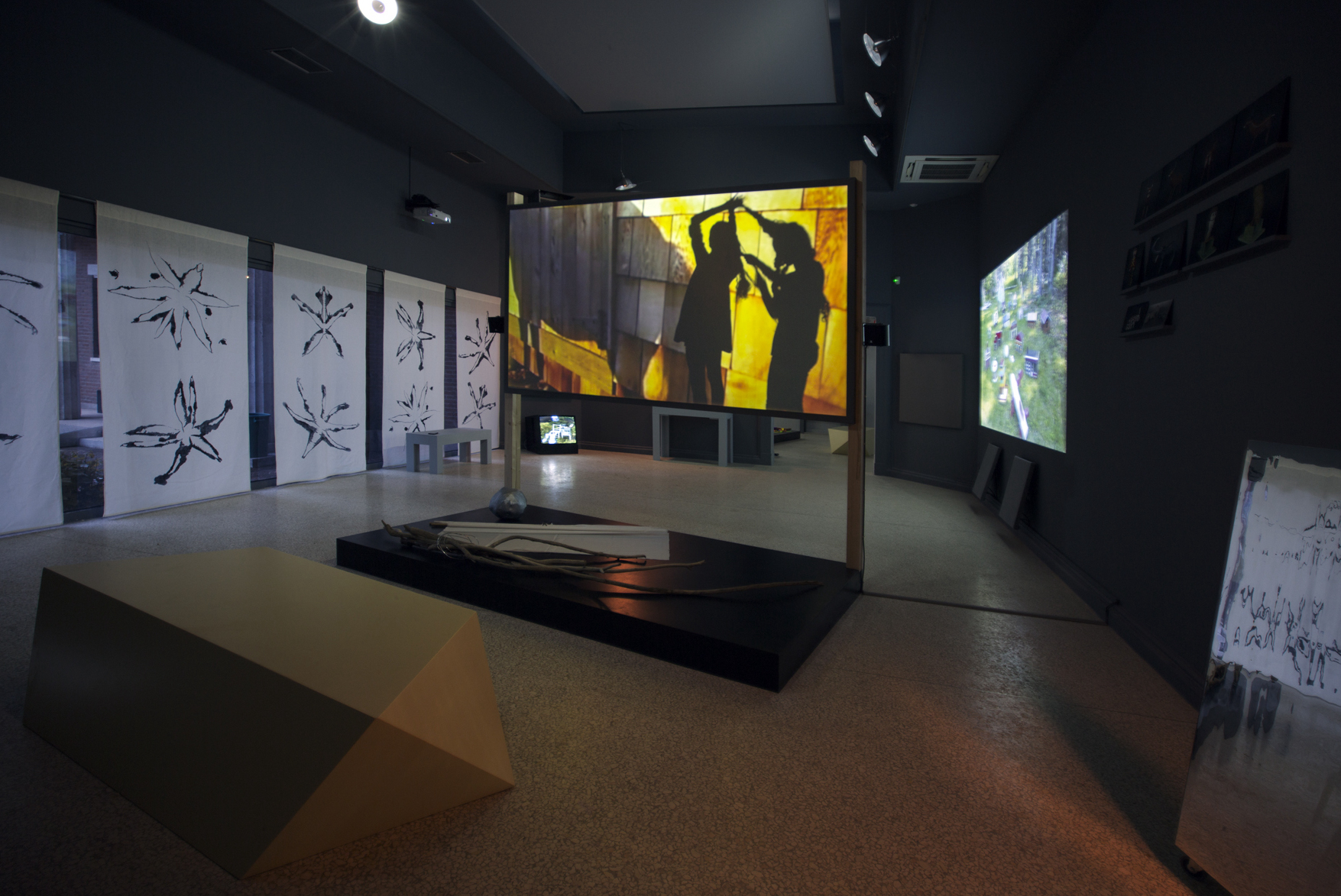 Installation view featuring a projector in the center of the room with two figure's shadows and prints lining the walls.