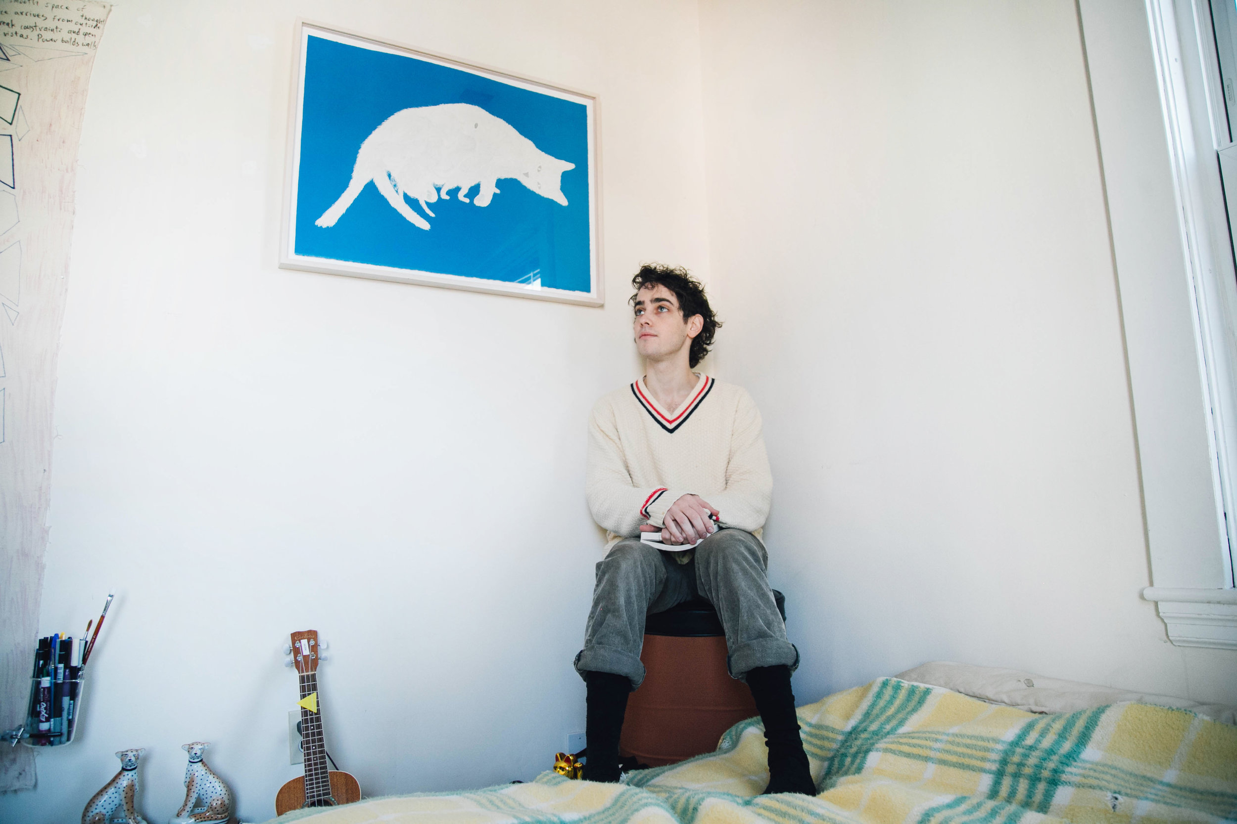 A man sits in the corner of his bedroom with a blue print on the wall behind him.