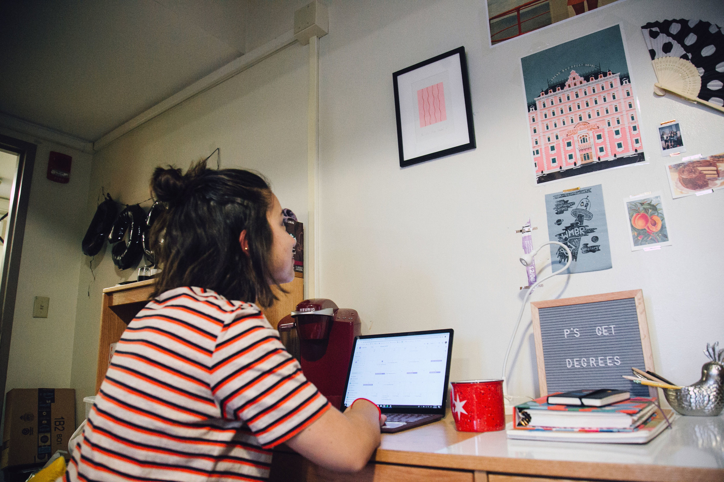 A girl in a red striped shirt sits at her dorm desk with her laptop open and gazes at the posters on the wall in front of her.