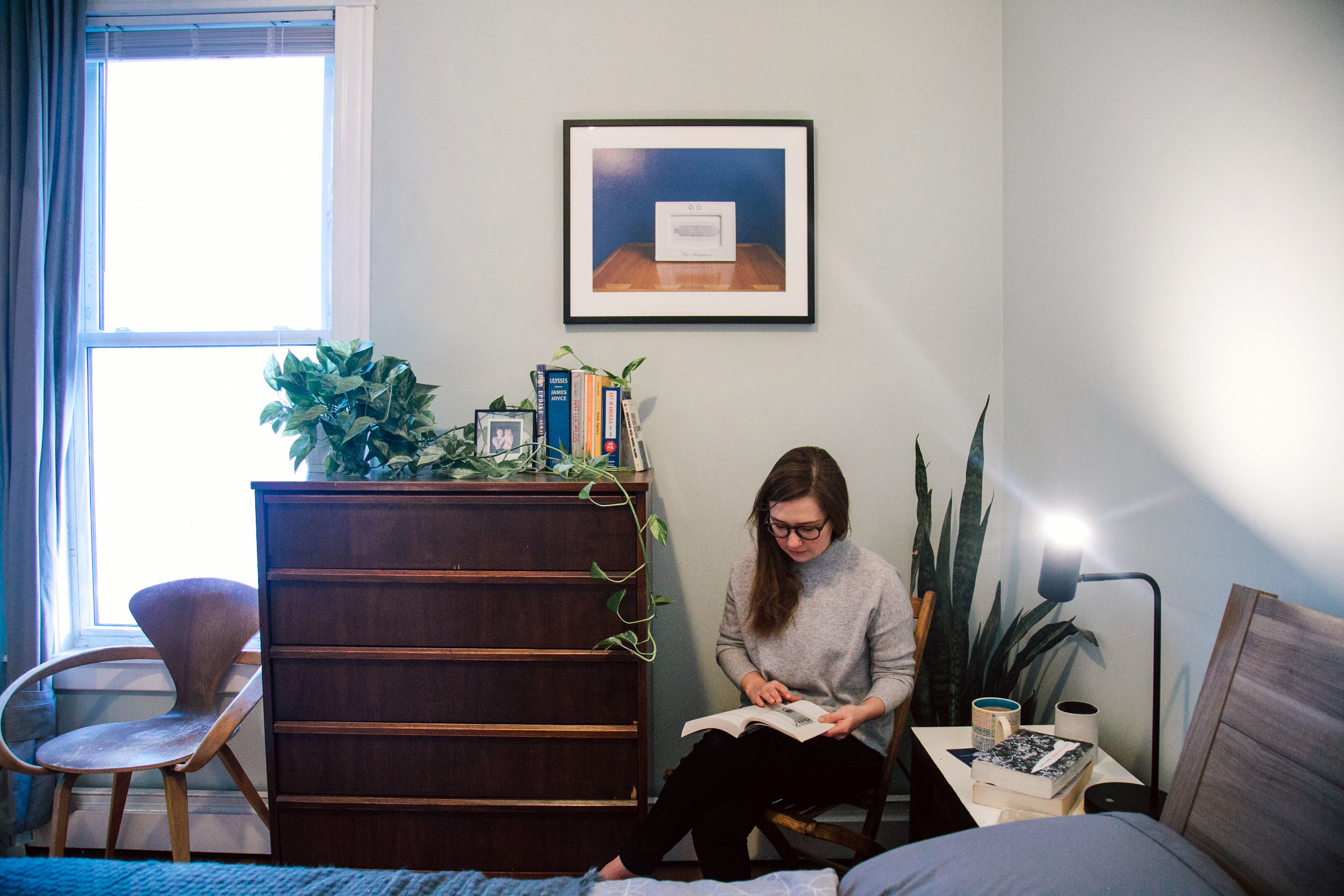A woman sits in the corner of her bedroom reading a book with a dresser to her right and a print on the wall behind her.