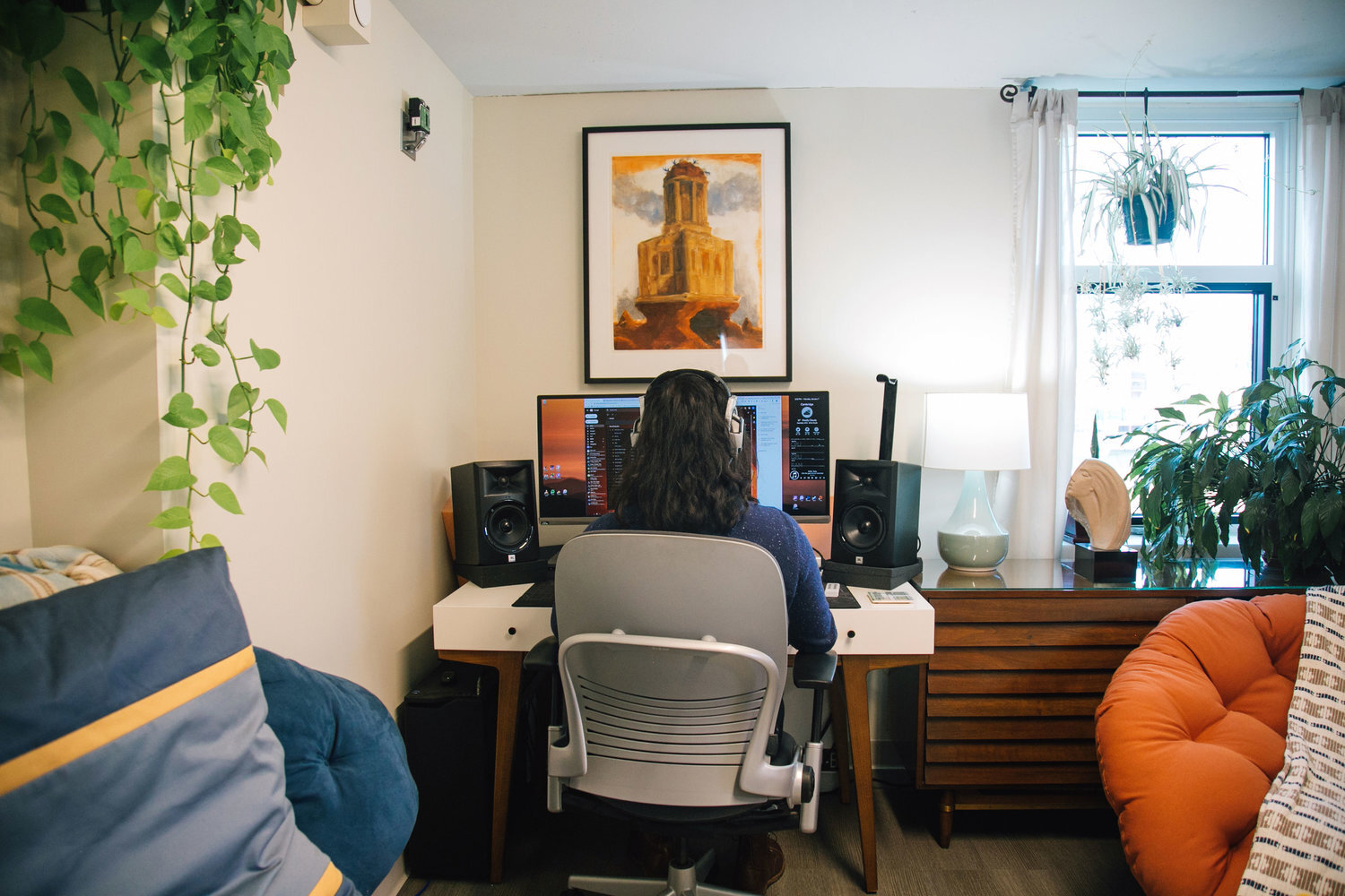 A man sits at his desk on his laptop surrounded by houseplants and a burnt orange mono print on the wall in front of him.
