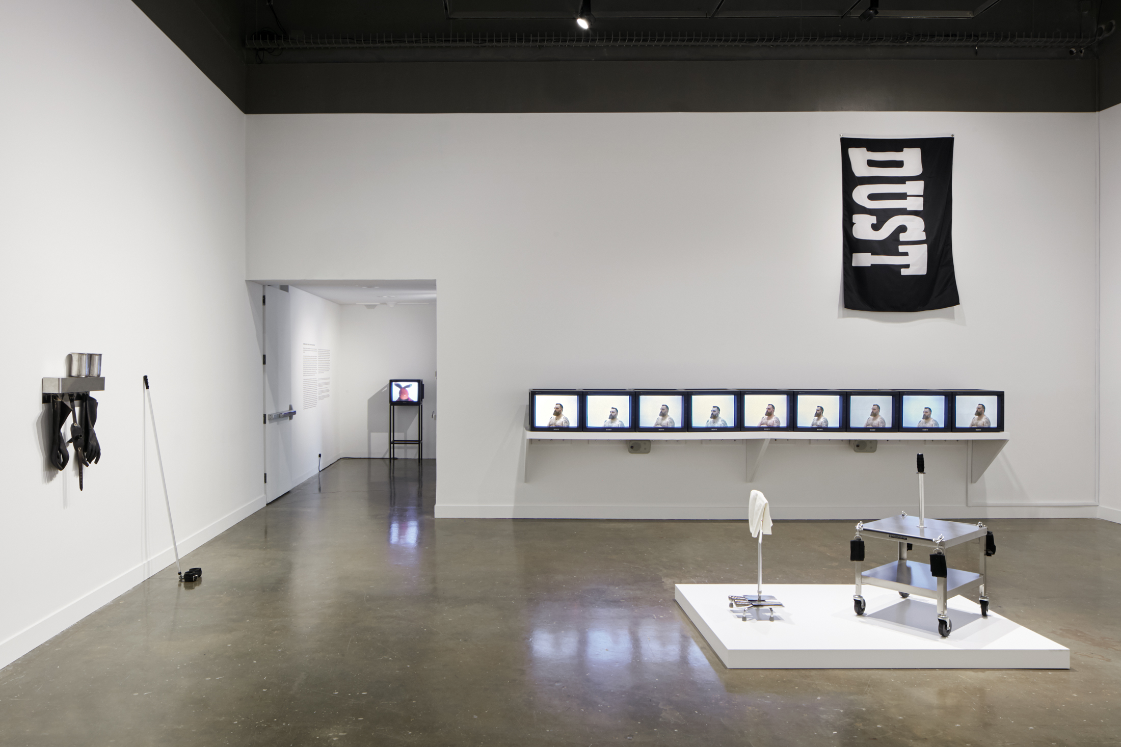 Installation v new featuring screens repeating in the center, janitor supplies hanging on the left wall and a metal sculpture in the center.