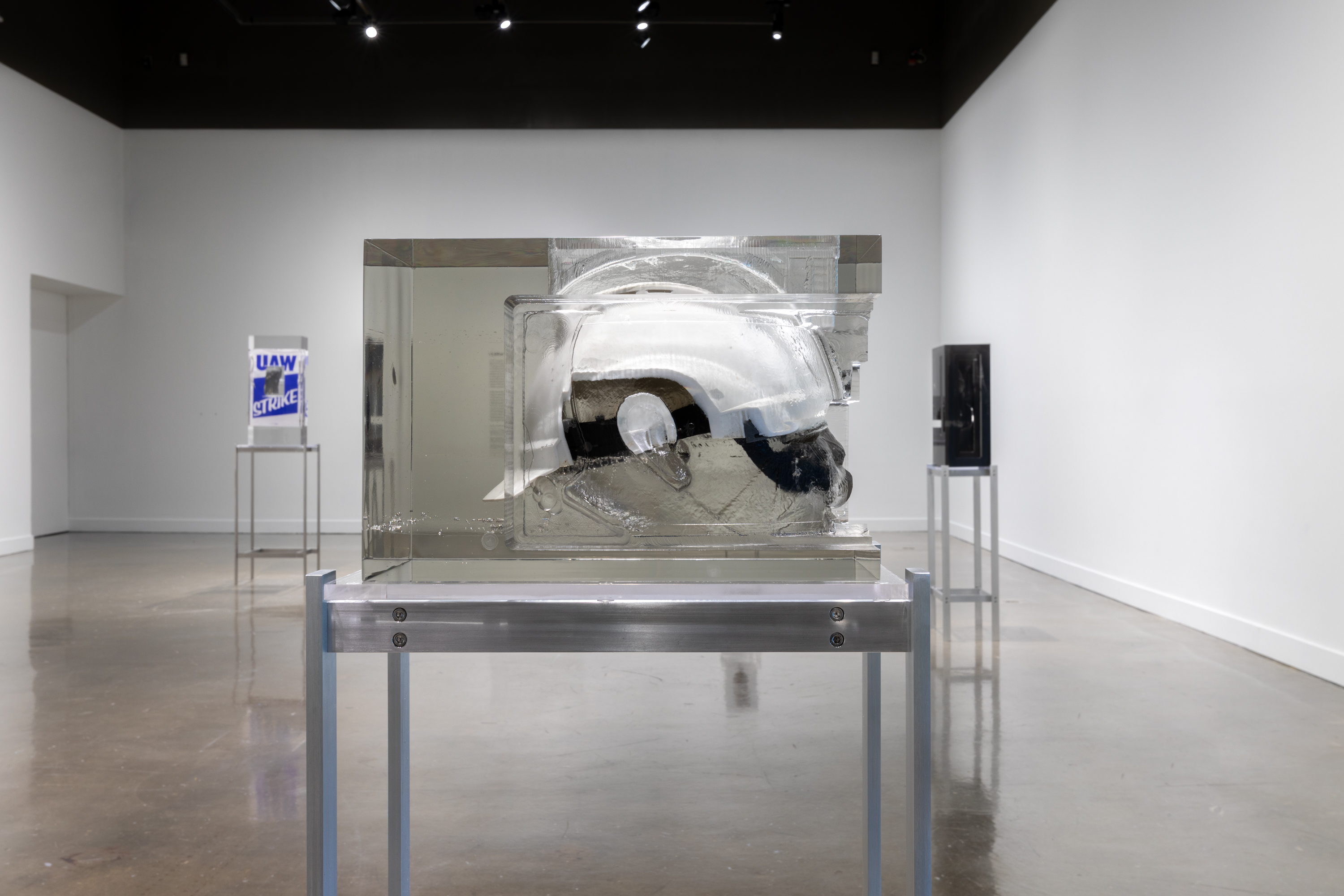 Installation view featuring a white helmet encased in a rectangular resin mold on a metal stand and two similar style sculptures in the background.