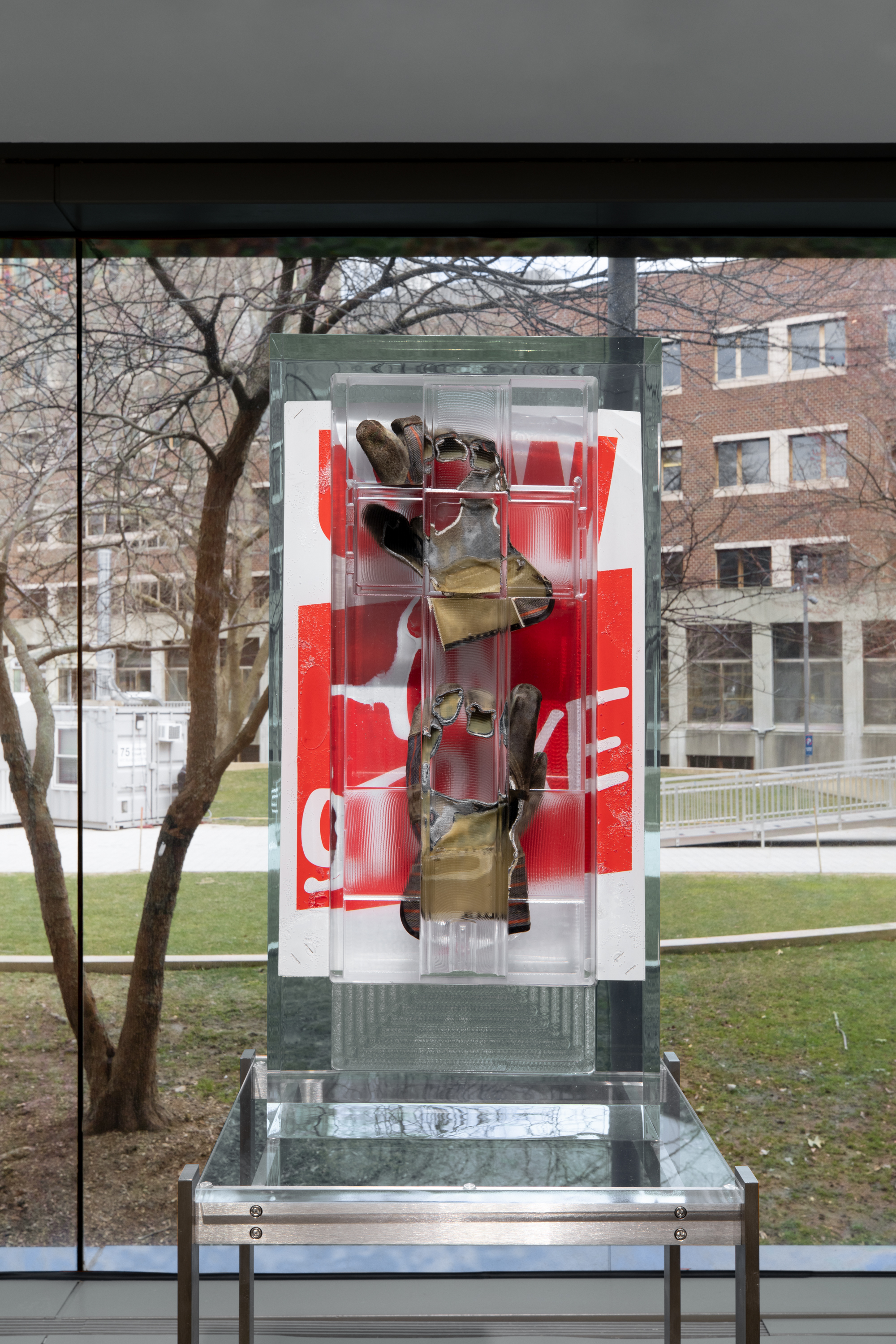 Installation view of a pair of work gloves and a red poster encased in a rectangular block of resin on a metal stand with a large window overlooking a courtyard in the background.