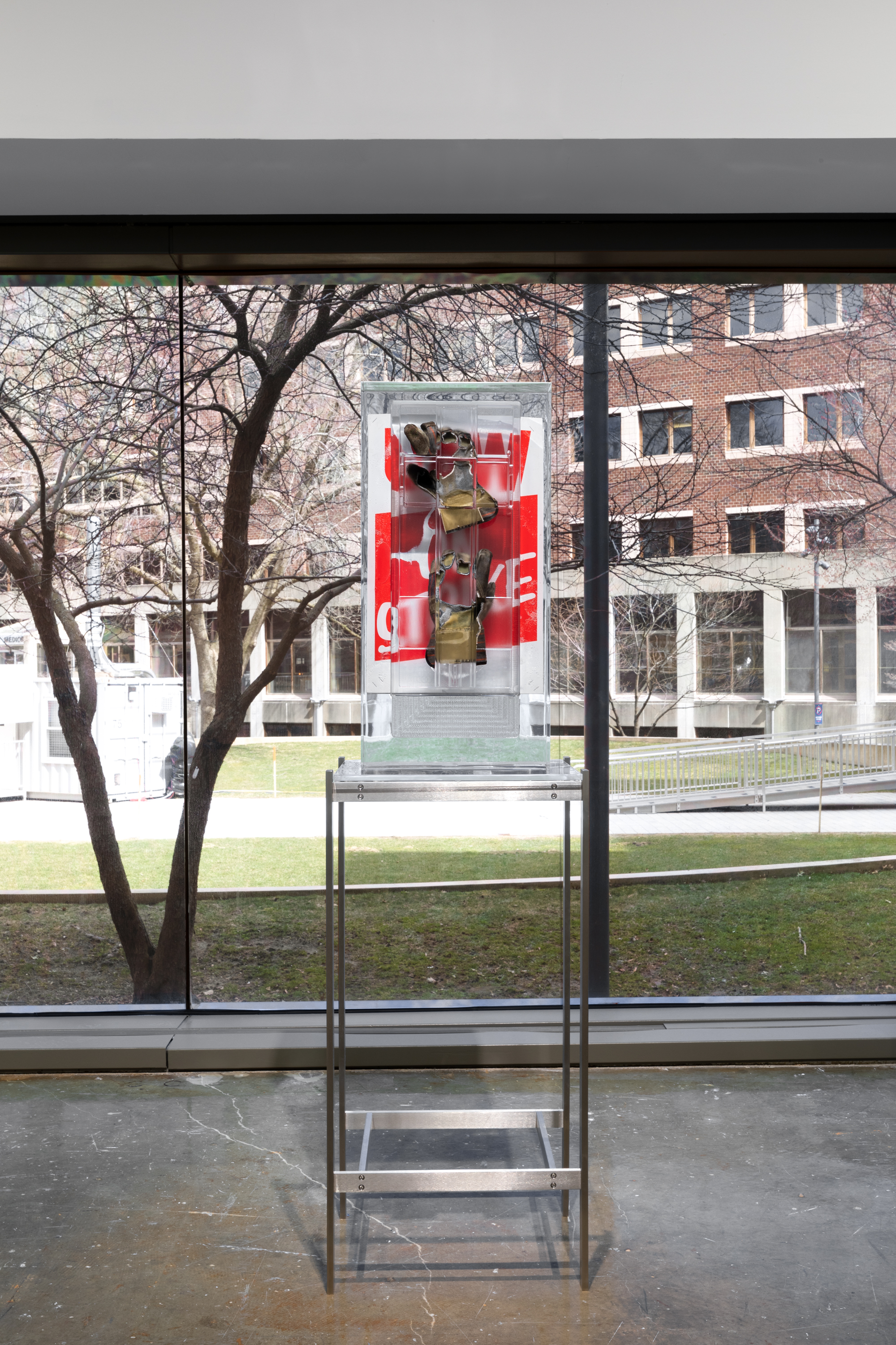 Installation view of a pair of work gloves and a red poster encased in a rectangular block of resin on a metal stand with a large window overlooking a courtyard in the background.