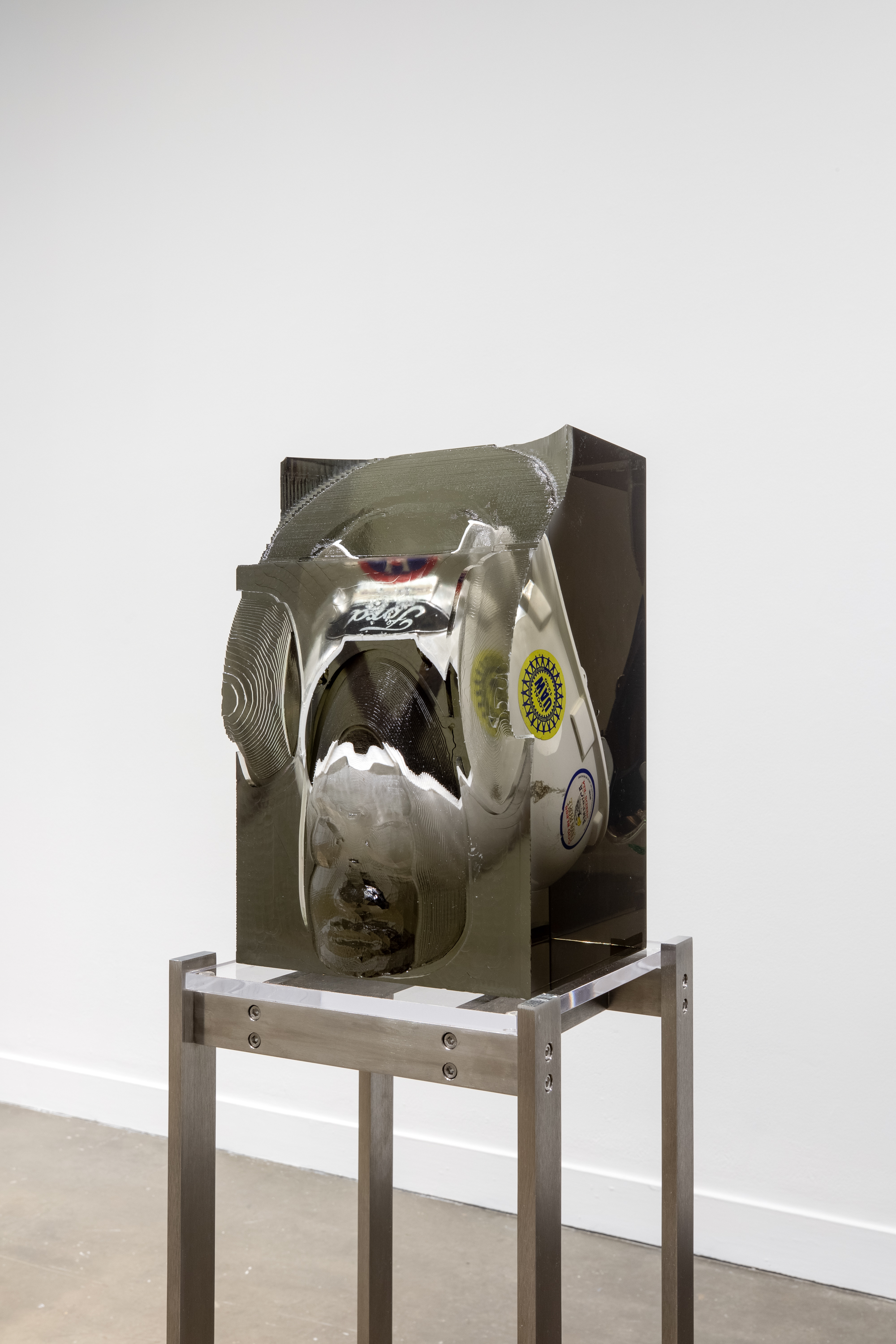 Installation view featuring a white Ford helmet with stickers on the side encased in a block of resin on a metal stand.