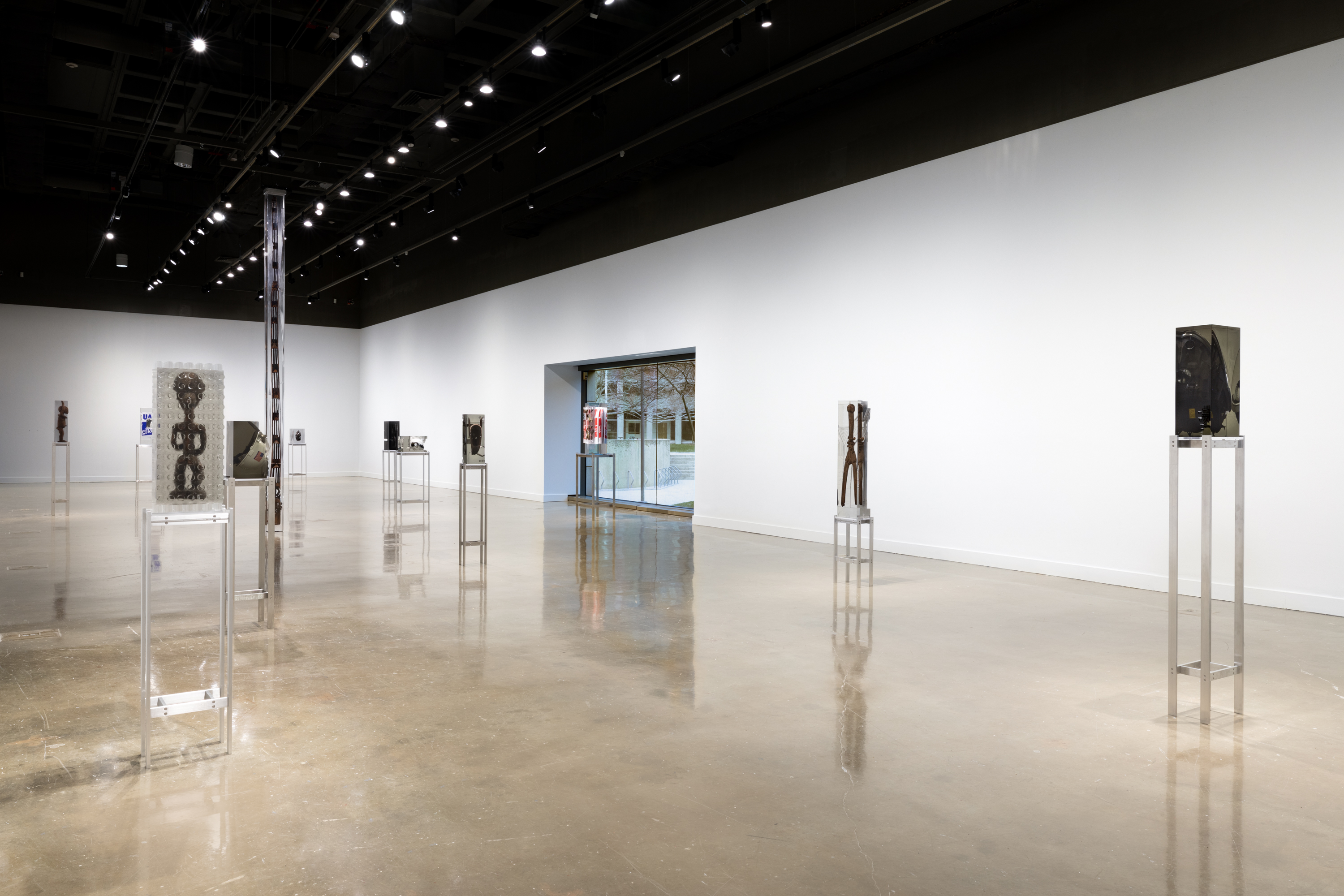 Exhibition view featuring 12 sculptures of varying sizes with objects inside of blocks of clear resin spread throughout the gallery. Each sculpture is on a tall metal stand.