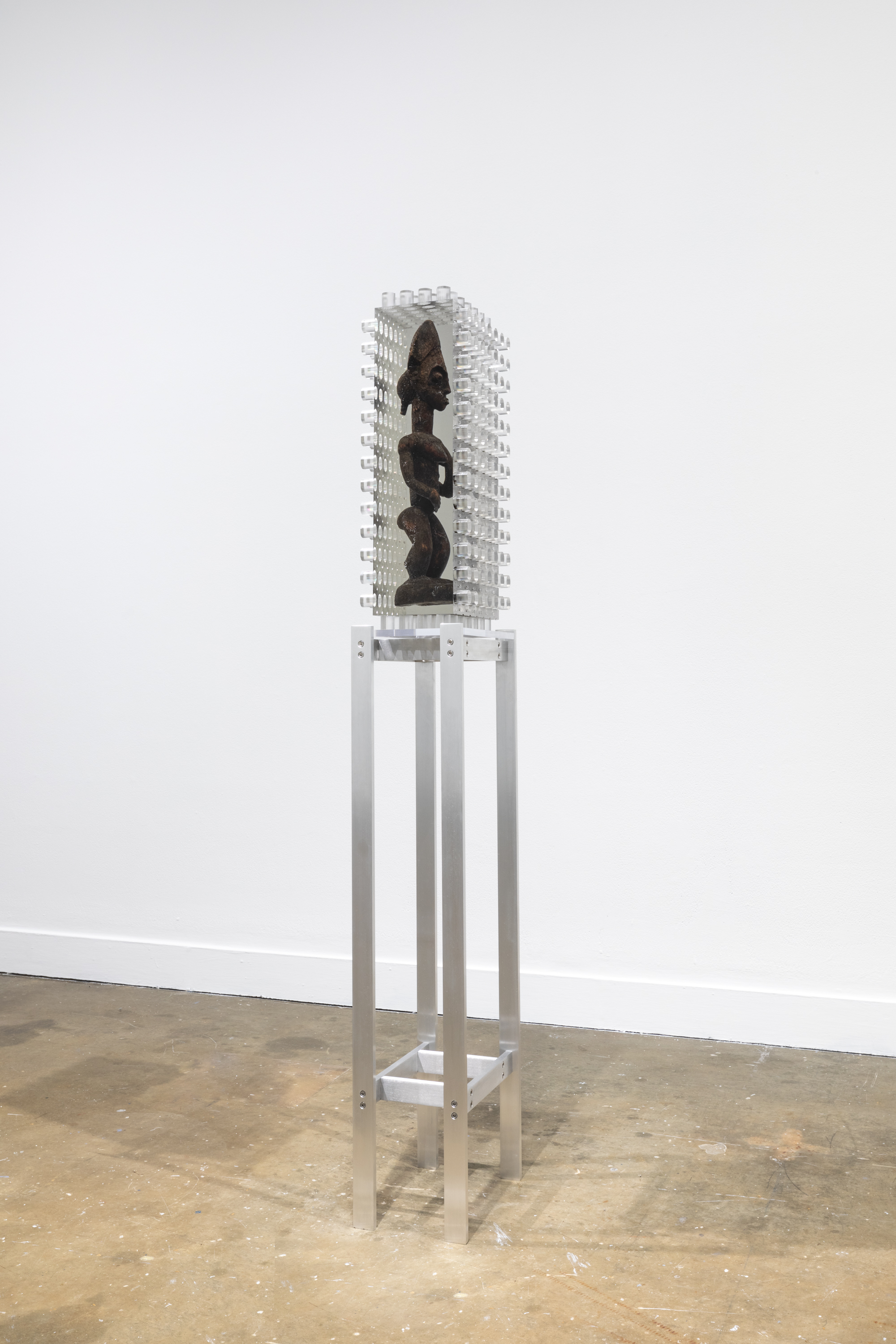 Installation view of an African wooden sculpture of a figure encased in a block of resin with jagged edges on a tall metal stand.